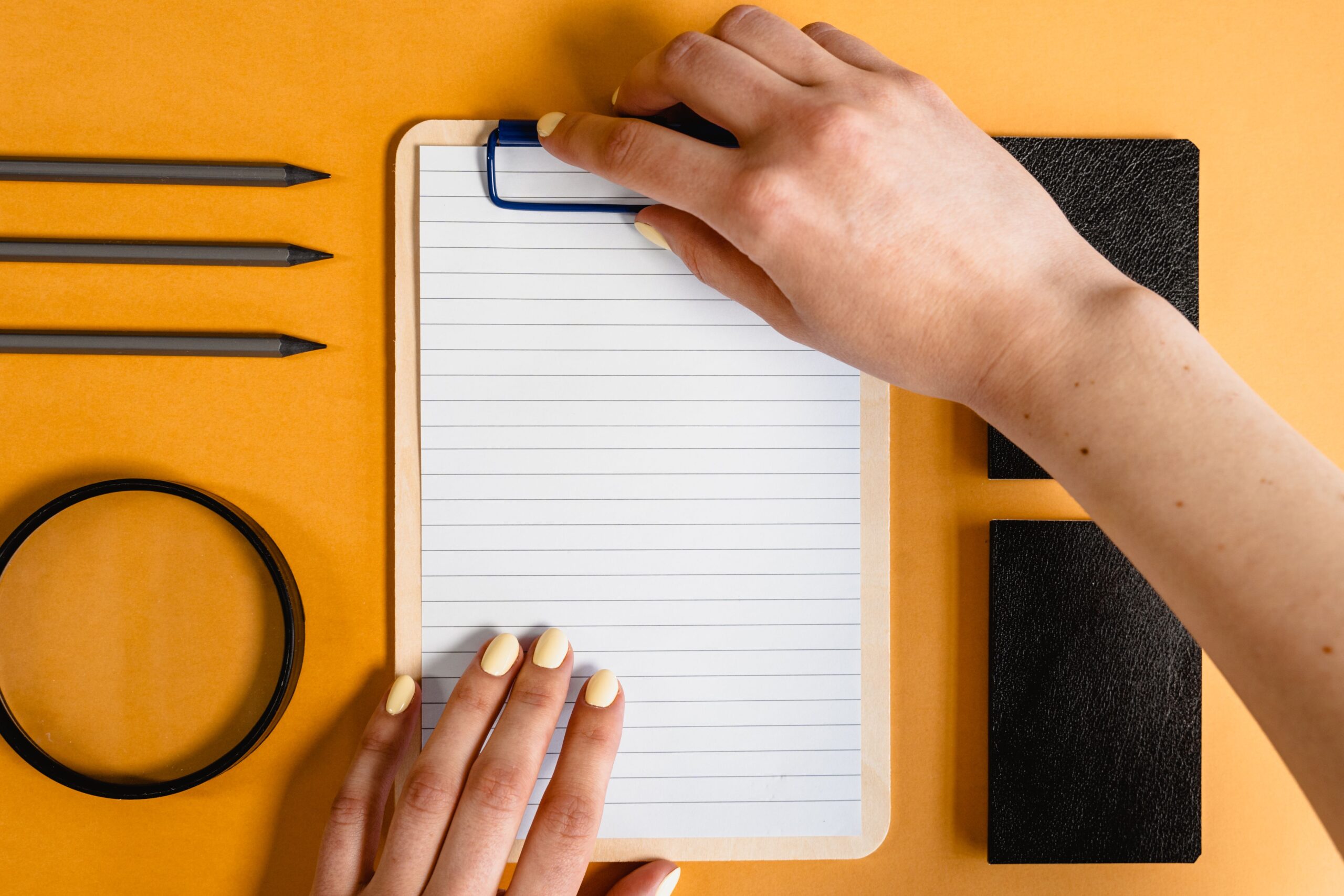 Top view of hands on a notepad with various stationery items on a yellow desk background.
